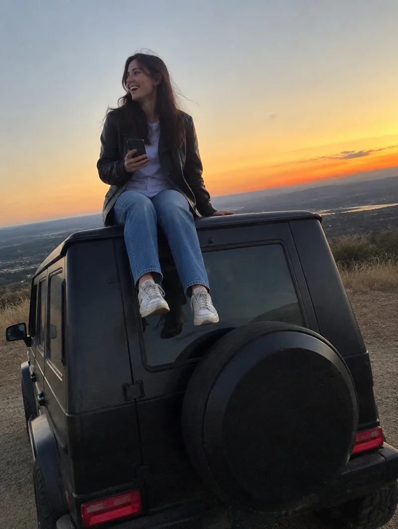 A woman sits on the roof of a black SUV above a valley at sunset, laughing, holding her phone.